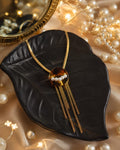 Gold-tone necklace with a round amber-brown centerpiece, white faux pearls, and three dangling chain fringe, displayed on a black leaf-shaped jewelry stand with warm pearl strands and fairy lights in the background.
